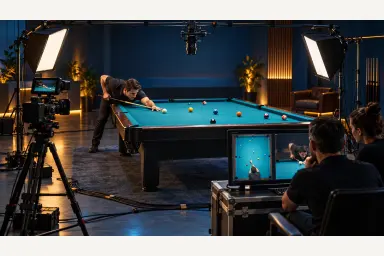 Illustration of a pool video crew framing an overhead table shot while a player prepares under soft studio lighting