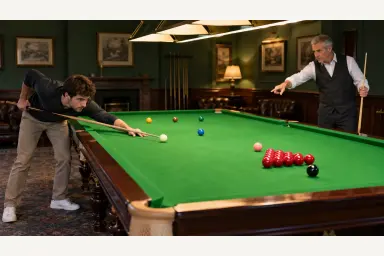 Beginner learning on a large green snooker table with red and colored balls under an instructor's guidance